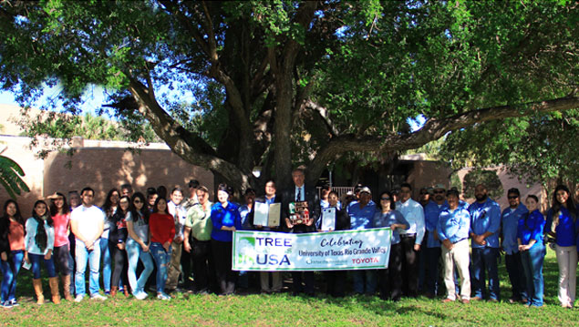 many UTRGV students, faculty, and staff infront of a big tree with people in the middle holding a tree campus usa designation banner
