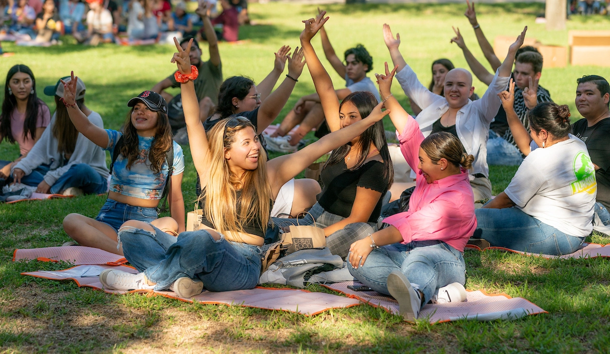 Students outdoors during event sitting on ground and signaling v's up while similing.