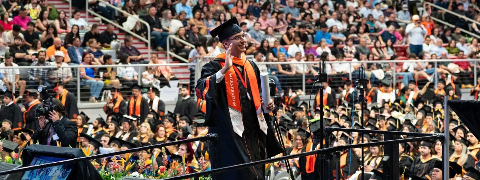 UTRGV graduate walking across the commencement stage while waving to the crowd inside a packed arena.