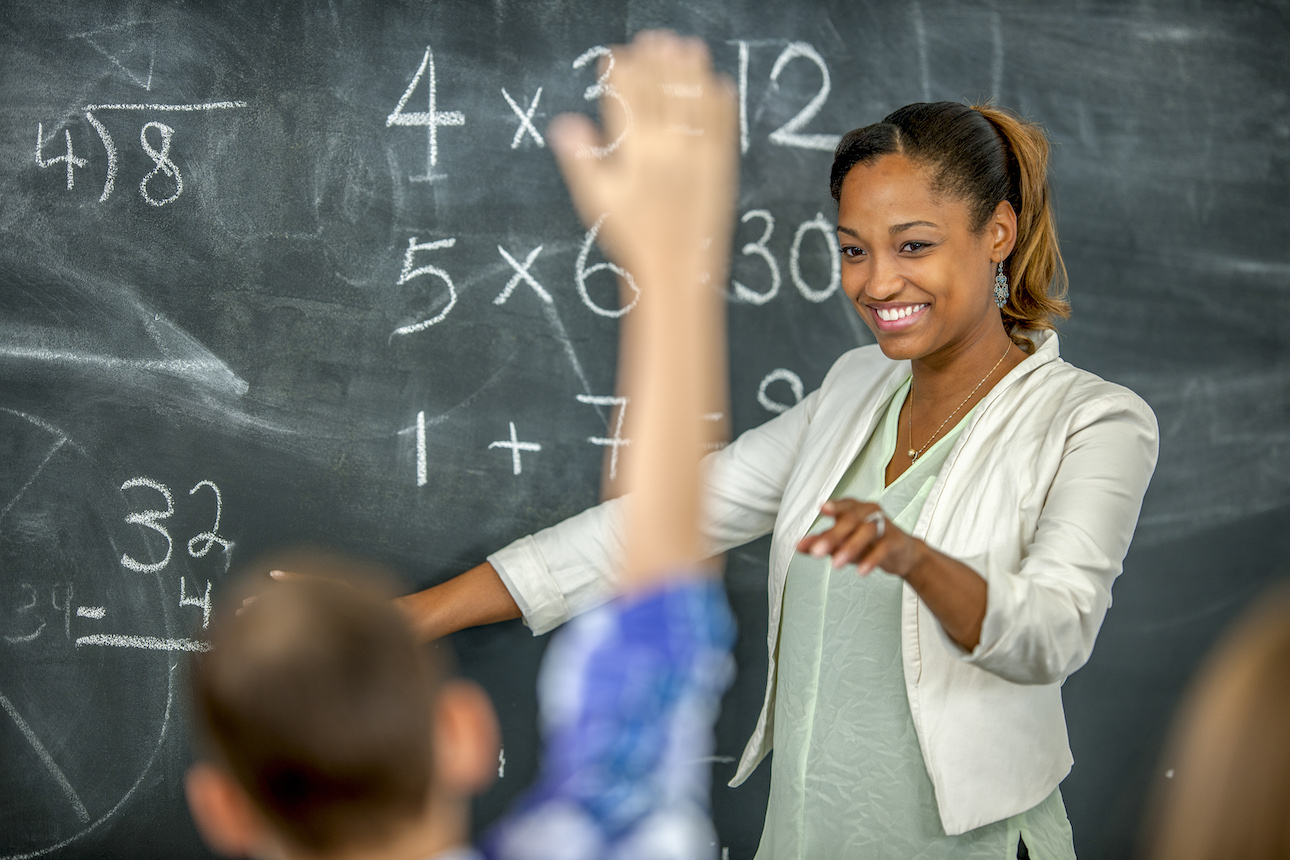 teacher at chalkboard pointing at student with hand raised