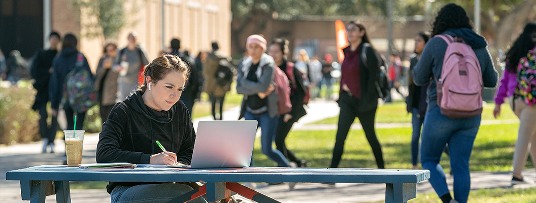 Female student on a picnic table, working on her laptop, enjoying an iced coffee drink