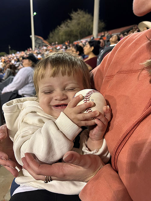 Child happily holding a baseball under her mother's arms.