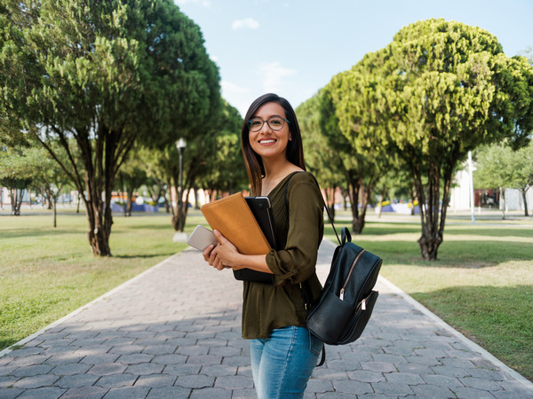 Smiling student holds books on campus walkway surrounded by trees