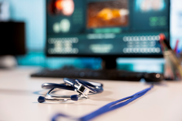A stethoscope and lanyard on a desk in front of a blurred medical computer screen.