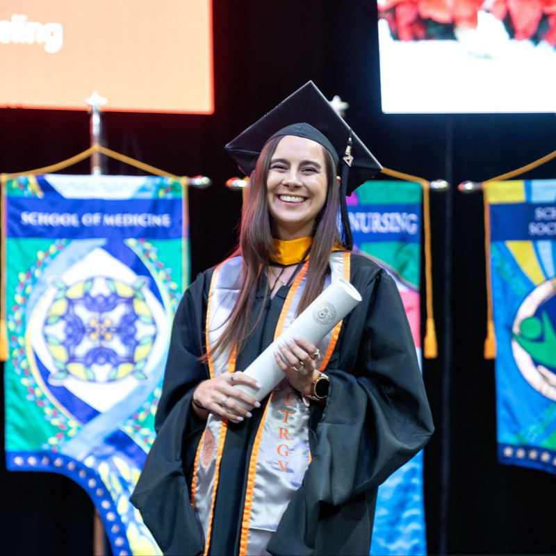 A smiling female graduate in a black cap and gown holding a diploma scroll on stage at a UTRGV commencement ceremony, with school banners in the background.