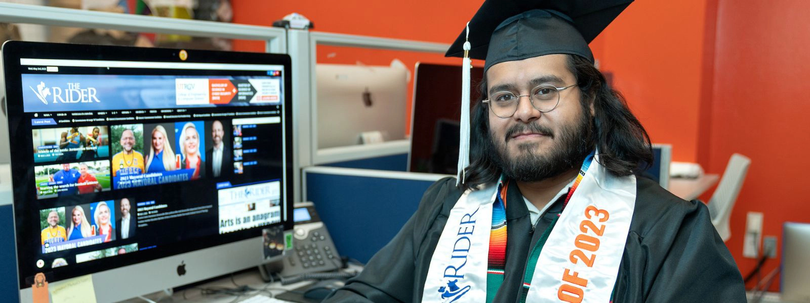 Student in cap and gown next to computer.