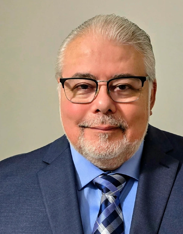Dr. René Gonzalez wearing a blue suit, light blue dress shirt, patterned blue tie, and black-framed glasses, smiling in a professional headshot against a neutral background.