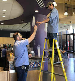 UTRGV facility employee giving a light bulb to another UTRGV facility employee on a ladder.
