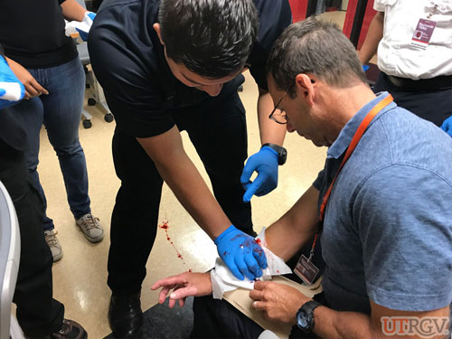 Sergeant Marco Rodriguez, wearing appropriate PPE, applies direct pressure with sterile gauze pads to stop the bleeding of a wound that EHSRM instructor John Guglielmo has simulated with stage blood.