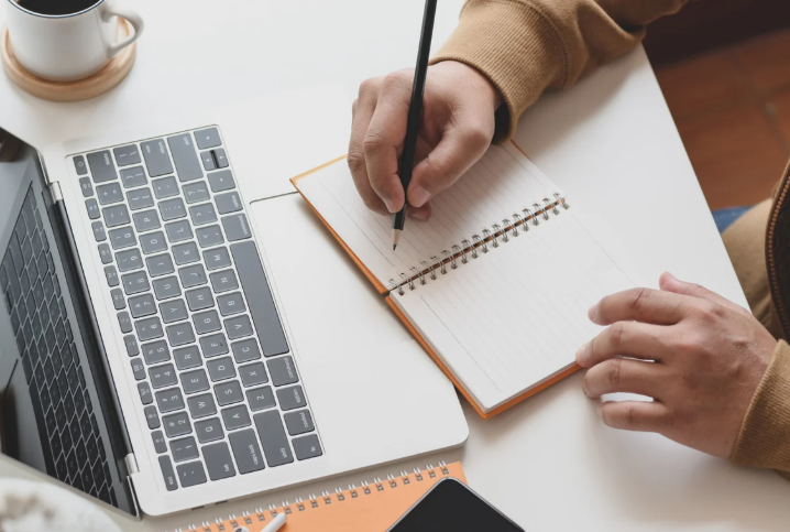 person writing on a notepad with a laptop open