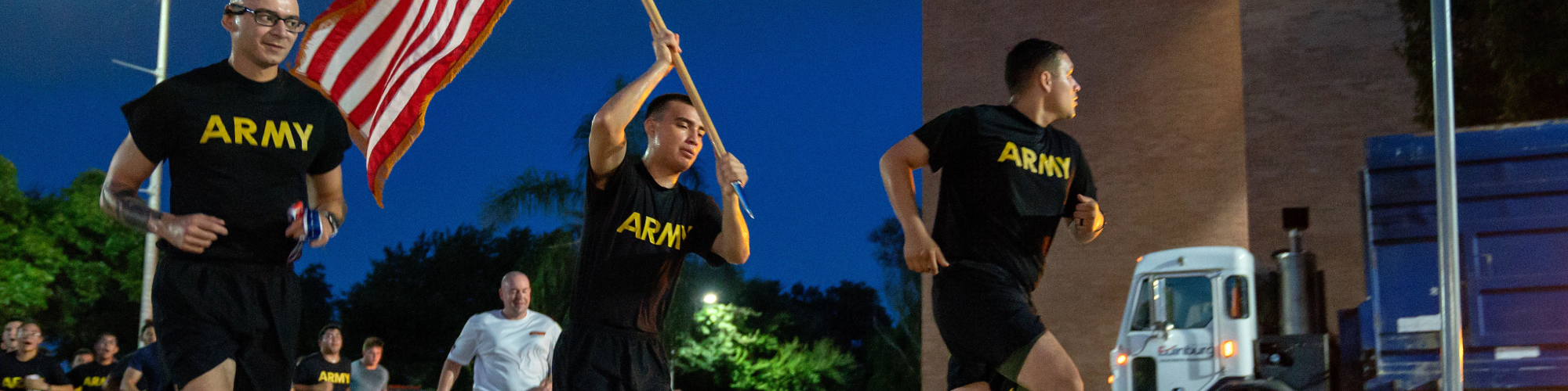 People running in race at night with t-shirts that say army and holding an American flag.