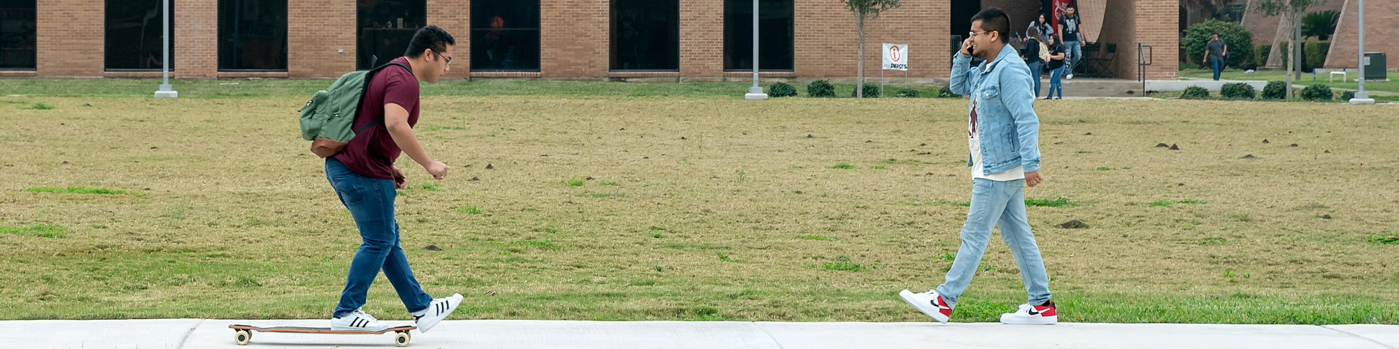 Student riding skateboard on campus and another student walking and talking on phone.