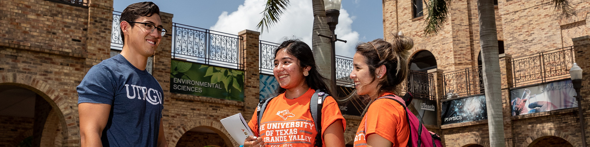 Three students wearing UTRGV shirts stand outside a brick campus building. Palm trees and a bright sky are in the background.