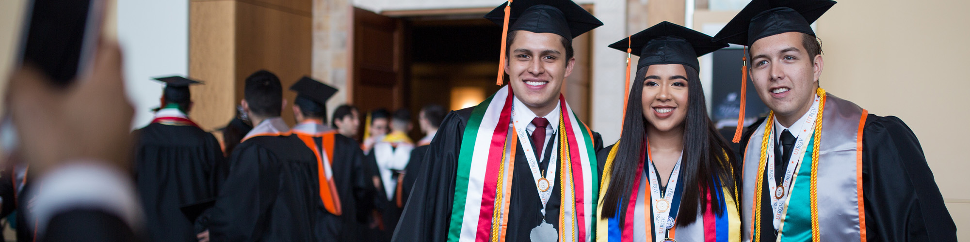 Three graduates in caps and gowns wearing colorful stoles and honor cords pose for a photo indoors, with other graduates walking in the background.
