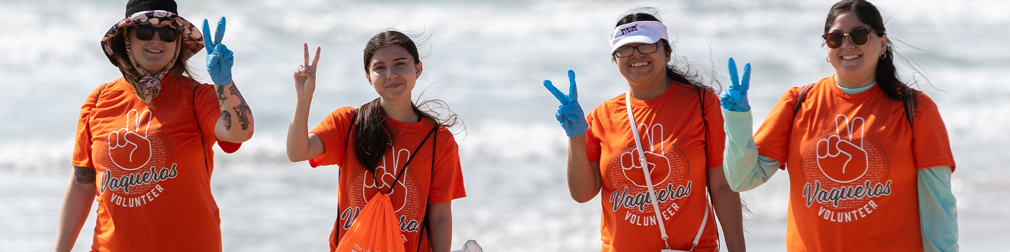 Four volunteers wearing bright orange ‘Vaqueros Volunteer’ T-shirts standing on a beach, holding up peace signs with their hands. Two of them are wearing blue gloves, and ocean waves are visible in the background.