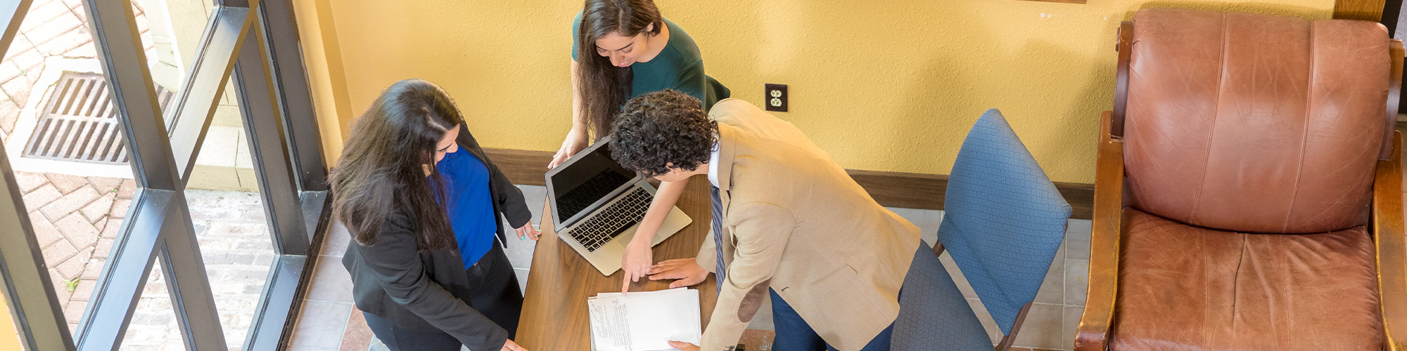 Three people collaborate at a wooden table with a laptop and papers in a bright office with large windows.