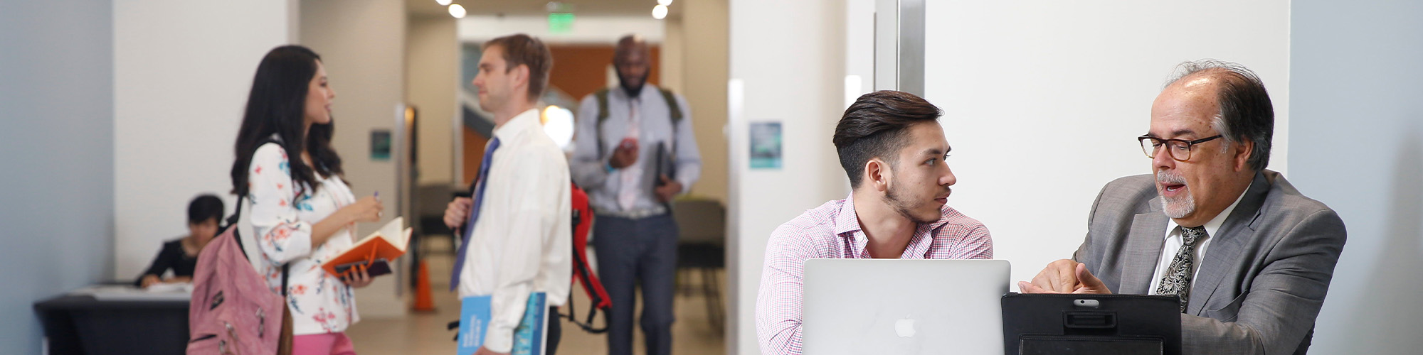 People interacting and working in a hallway area; two individuals seated at a table with laptops, while others stand and talk nearby.