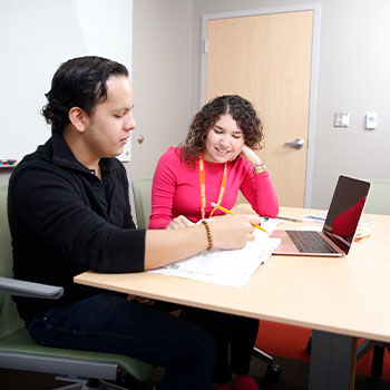 Two individuals seated at a table collaborating on paperwork with a laptop open nearby.