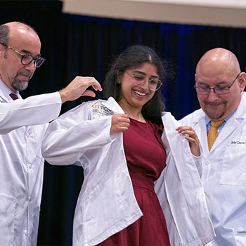 Medical school ceremony where a student is being assisted in putting on a white coat by two faculty members.
