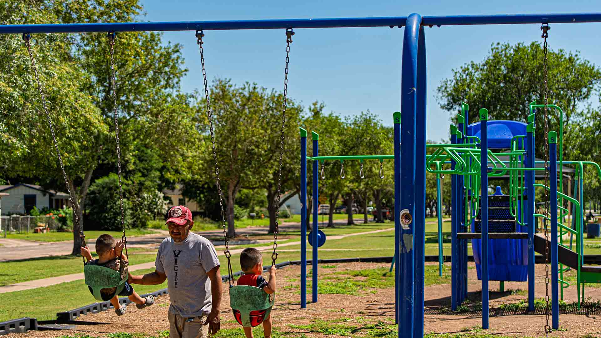 Adult pushes children on playground swings at a neighborhood park, supporting family connection, play, and healthy community living.