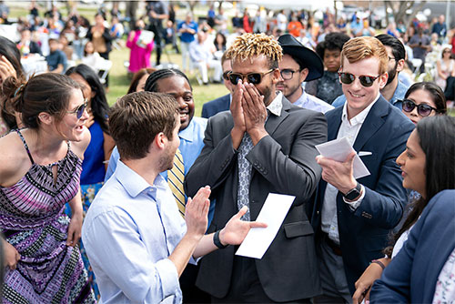 Group of people huddle around center male student, who is excited with joy in disbelief