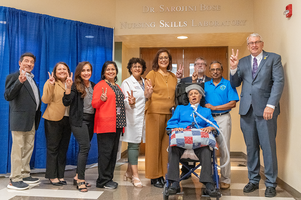 A group of people pose together in front of the “Dr. Sarojini Bose Nursing Skills Laboratory” sign, with one person seated in a wheelchair at the front.