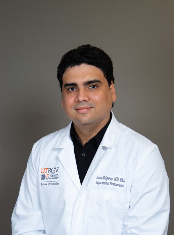 A person wearing a white UTRGV School of Medicine lab coat over a black shirt is posed against a plain gray background. The lab coat includes embroidered text for the School of Medicine and the Department of Neuroscience.