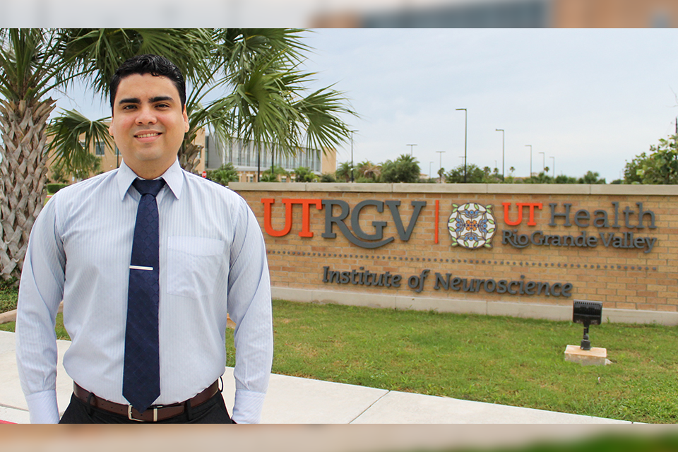 A person in professional attire stands outdoors in front of a brick monument sign that reads ‘UTRGV | UT Health Rio Grande Valley – Institute of Neuroscience,’ with campus buildings, palm trees, and grassy landscaping in the background.