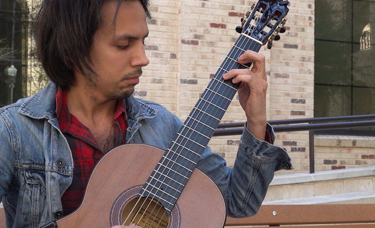 A person seated on an outdoor bench plays an acoustic guitar, wearing a denim jacket over a red plaid shirt, with a brick building in the background.