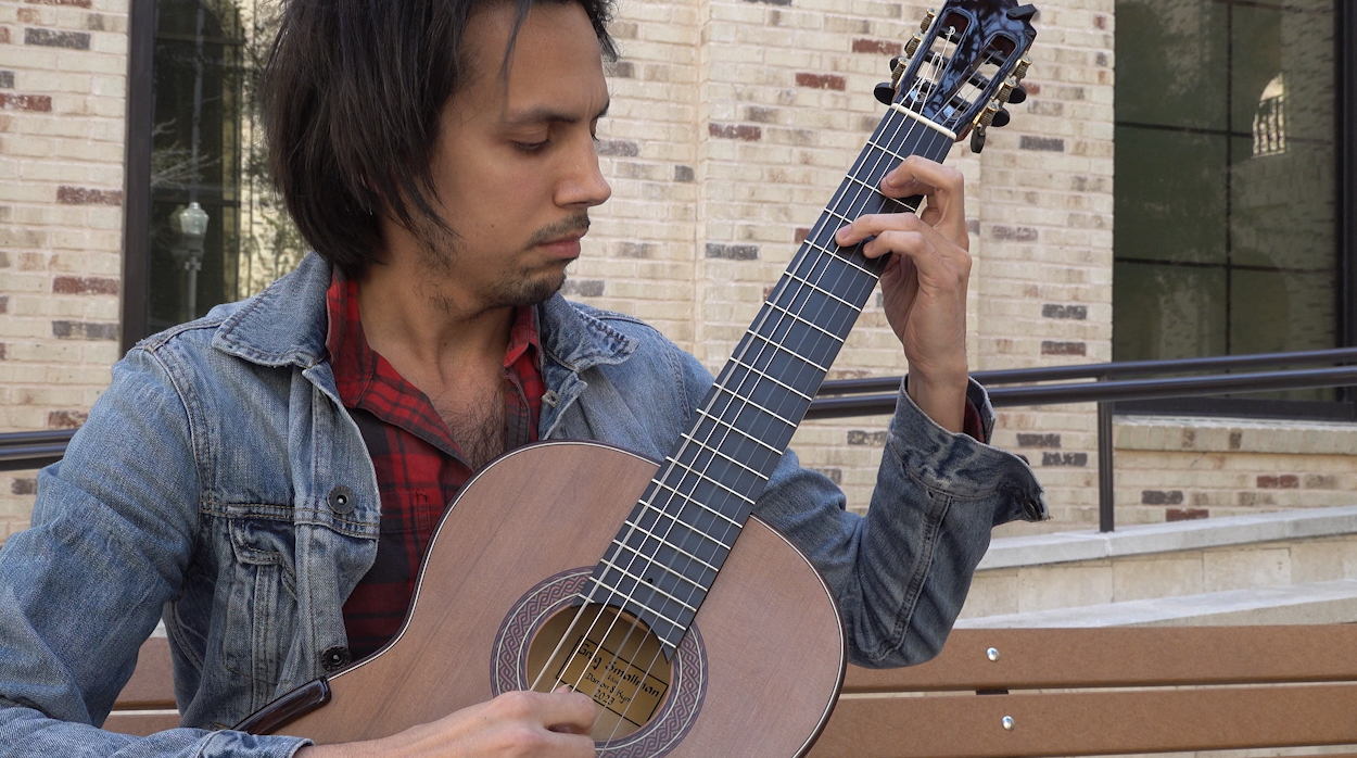 A person seated on an outdoor bench plays an acoustic guitar, wearing a denim jacket over a red plaid shirt, with a brick building in the background.