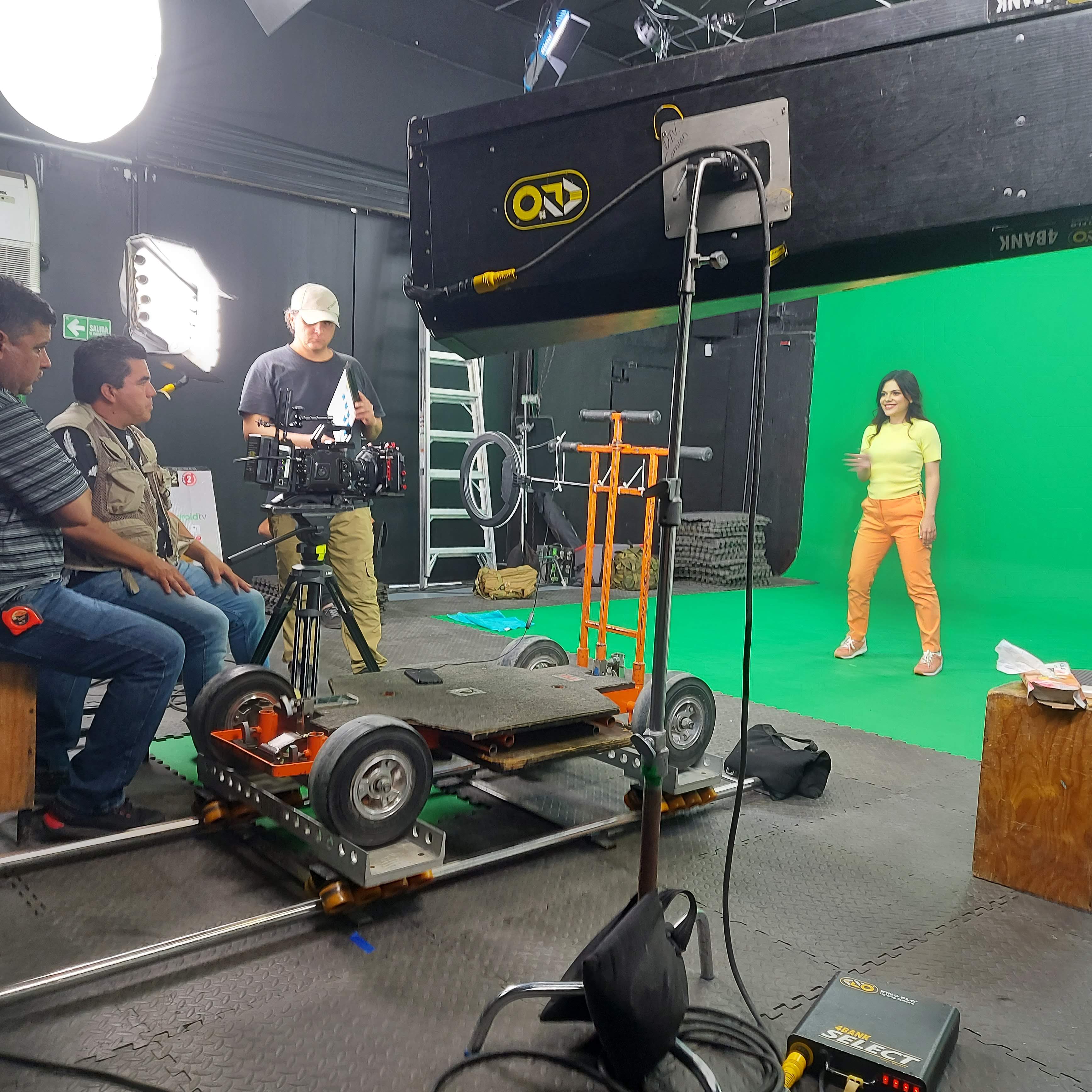 A video production crew works inside a studio equipped with cameras, lights, and a dolly track, while a performer stands in front of a large green screen.