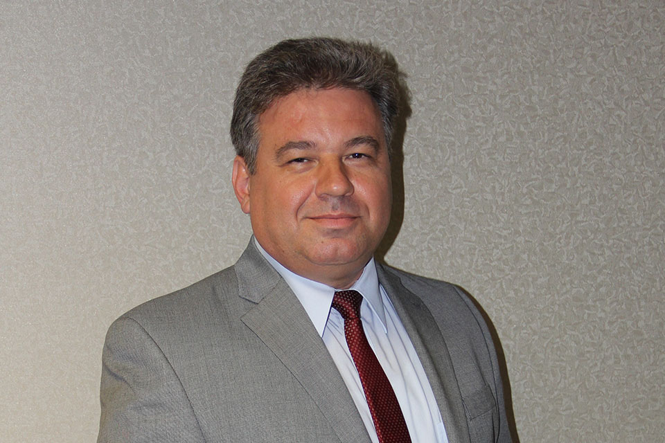 “A person wearing a light gray suit jacket, white dress shirt, and patterned red tie stands in front of a textured beige wall.