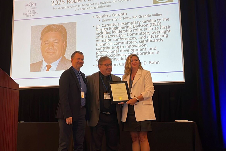Three individuals stand on a stage holding a framed certificate in front of a large presentation screen. The screen displays text describing an engineering award and includes a headshot and organizational logos.