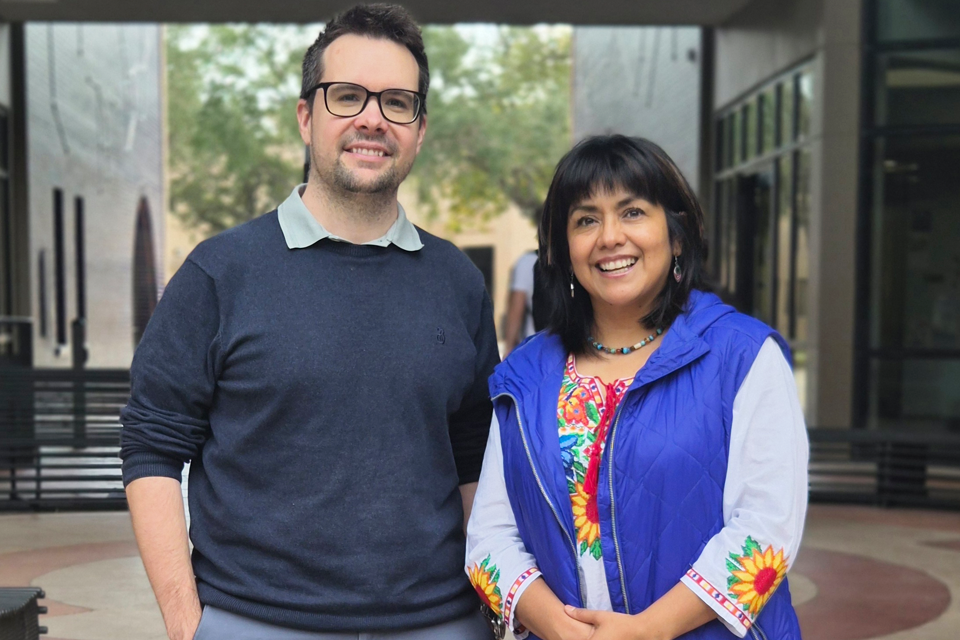 Two people stand side by side in an outdoor courtyard area, with modern building structures and trees in the background. One person is wearing a dark long-sleeve sweater, and the other is wearing a colorful embroidered top with a blue vest. They are positioned in the center of the image, facing the camera.