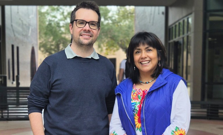 Two people stand side by side in an outdoor courtyard area, with modern building structures and trees in the background. One person is wearing a dark long-sleeve sweater, and the other is wearing a colorful embroidered top with a blue vest. They are positioned in the center of the image, facing the camera.