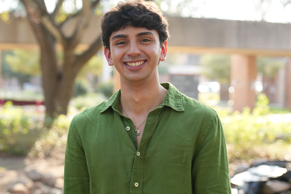A person wearing a green button-up shirt stands outdoors in a sunlit area with trees and a concrete walkway in the background.