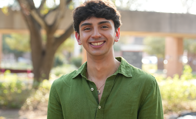 A person wearing a green button-up shirt stands outdoors in a sunlit area with trees and a concrete walkway in the background.