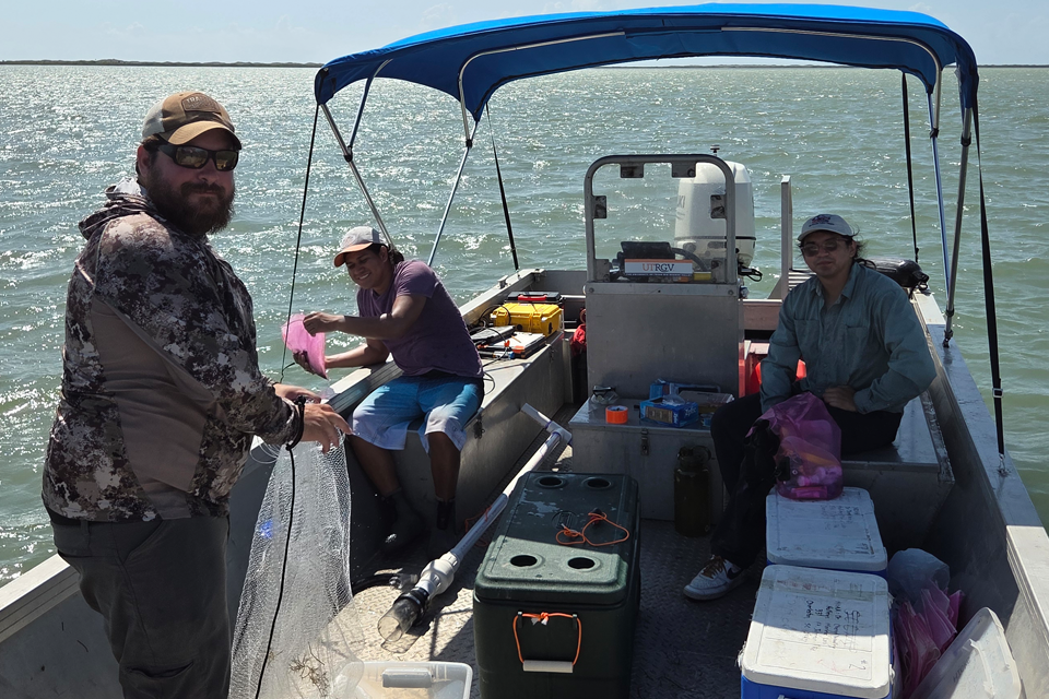 A group of people are on a small aluminum boat on open water. One person stands at the left holding a fishing net, while another sits near the center handling a pink cloth or bag. A third person sits on the right side with gear beside them. The boat contains various equipment, including coolers, ropes, containers, and tools. Sunlight reflects off the water in the background.