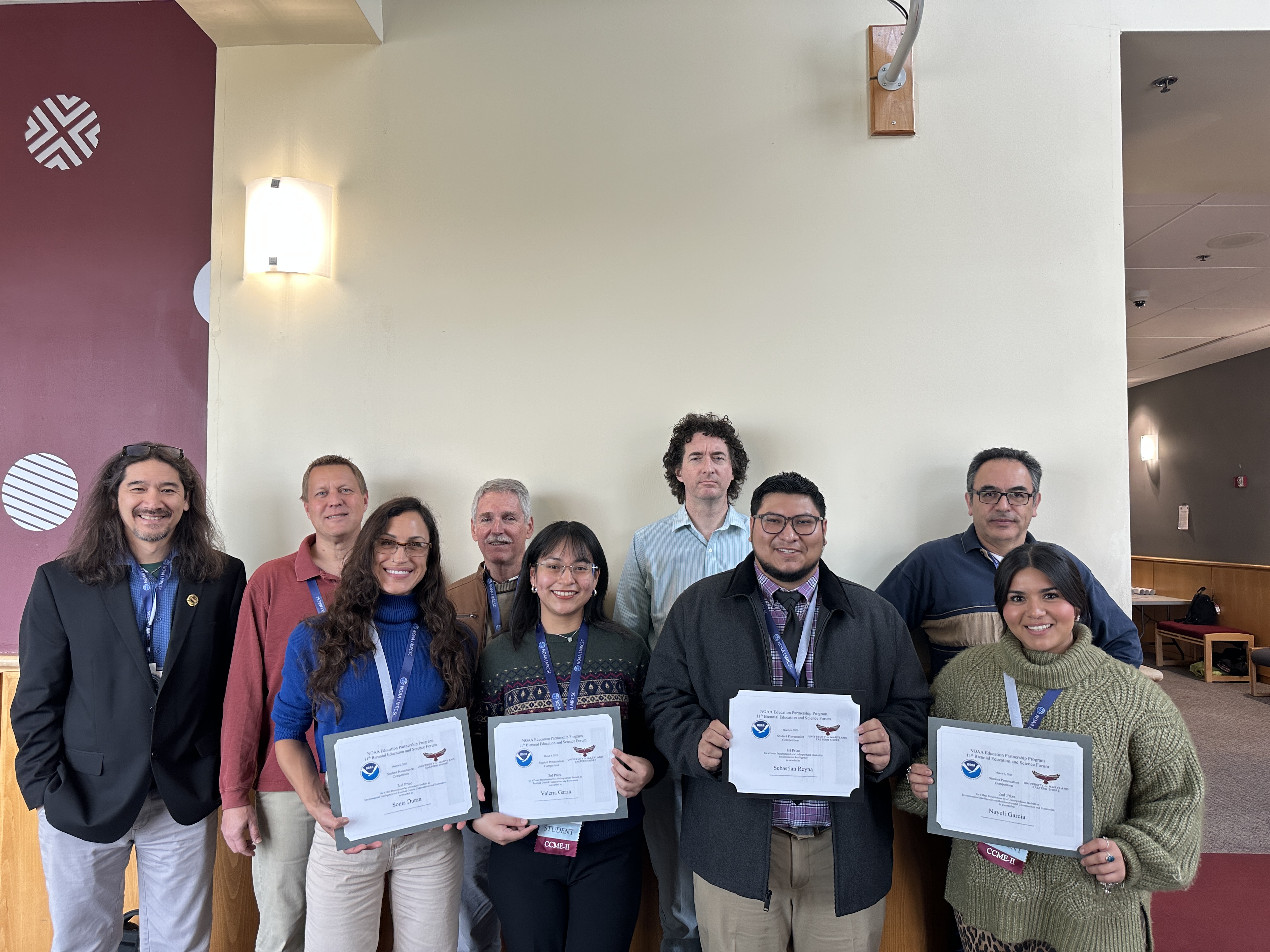 A group of nine people pose indoors against a light‑colored wall. Four people in the front row hold certificates featuring NOAA logos. Some individuals wear conference badges around their necks. The group stands in a lobby or meeting space with neutral‑colored walls, wood paneling, and overhead lighting.