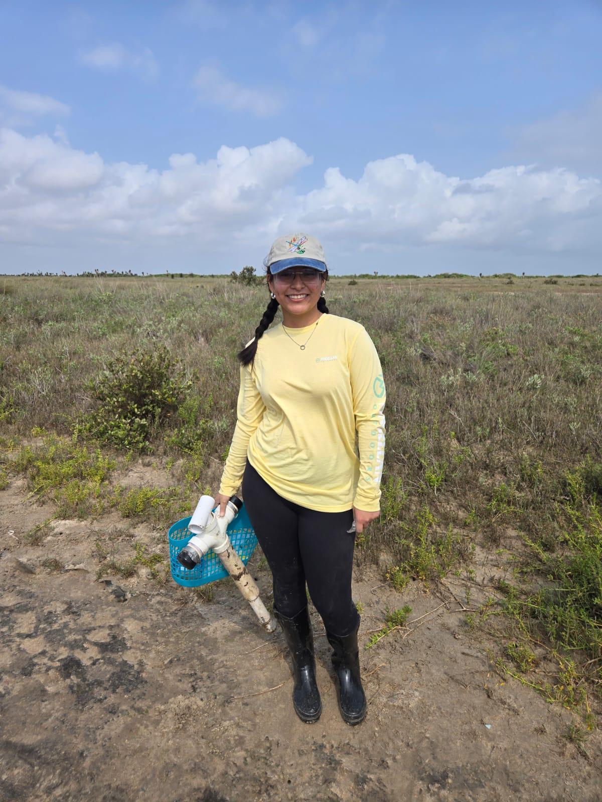 A person stands outdoors in an open grassy field under a partly cloudy sky. The person is wearing a long‑sleeved yellow shirt, dark pants, rubber boots, and a cap. They are holding a blue plastic basket and a piece of white PVC pipe with attachments. Low shrubs and natural vegetation surround the dirt path where they stand.