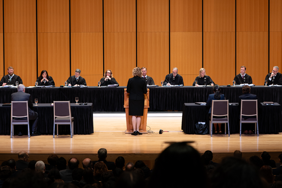 A large auditorium filled with seated attendees faces a stage where a panel of judges in black robes stands behind long tables arranged in a semicircle. The space features tall wooden acoustic panels, multiple cameras on tripods, and U.S. and Texas flags positioned on either side of the stage.