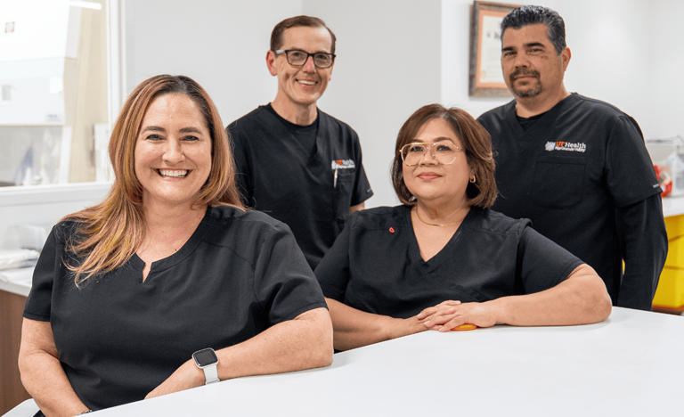 Four healthcare professionals in black scrubs stand and sit confidently around a white counter in a bright, modern clinic, expressing teamwork and professionalism.