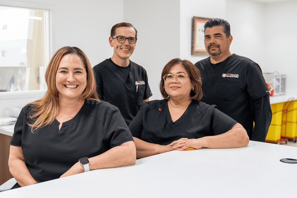 Four healthcare professionals in black scrubs stand and sit confidently around a white counter in a bright, modern clinic, expressing teamwork and professionalism.
