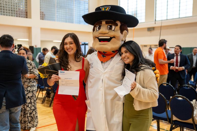 Two women smiling with a mascot in a white coat at an indoor event. One holds a bouquet and document, the other also has a document. Joyful atmosphere.