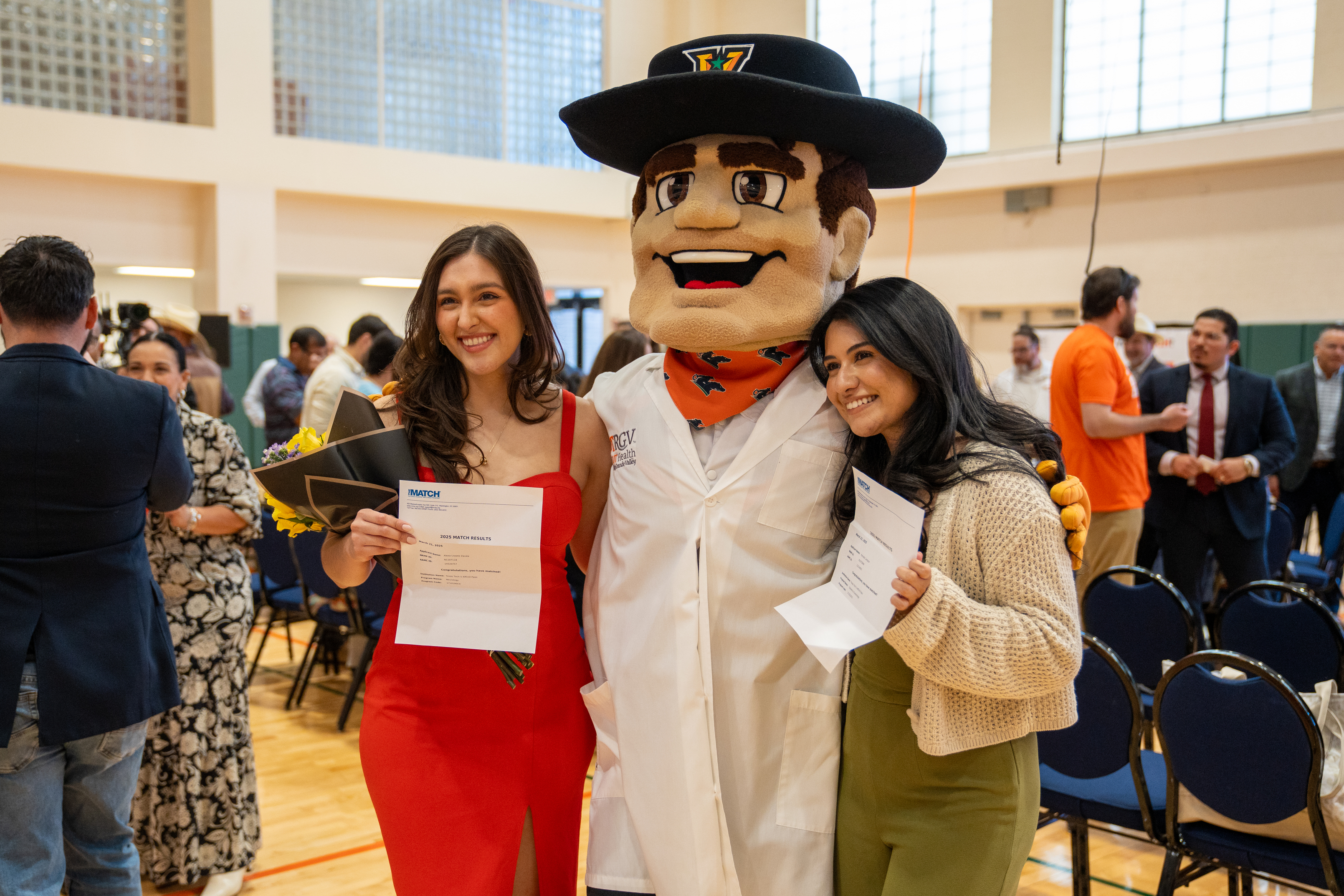 Two women smiling with a mascot in a white coat at an indoor event. One holds a bouquet and document, the other also has a document. Joyful atmosphere.
