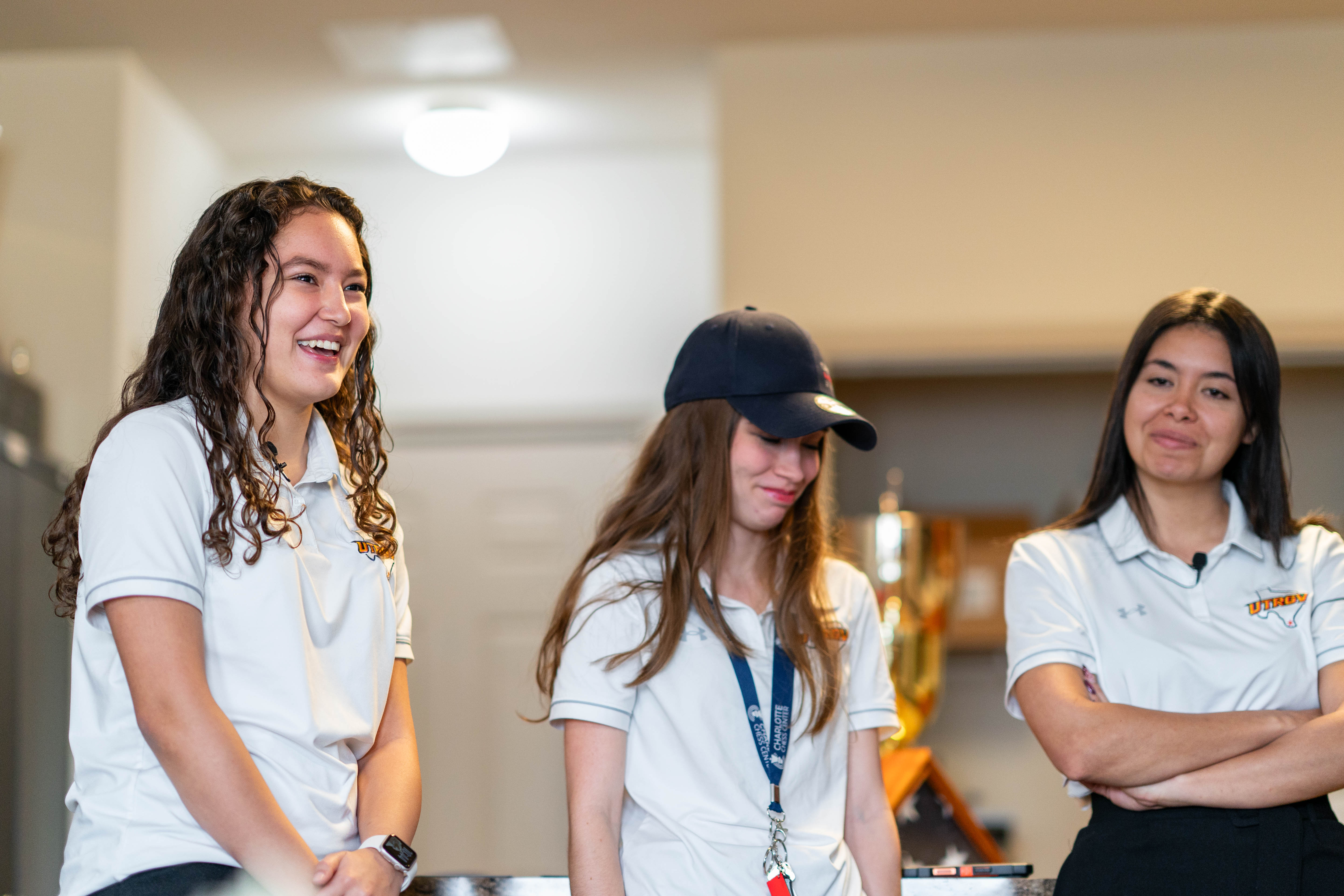 Three women wearing a white polo Chess shirts, as they stand in an room with trophies in the background.