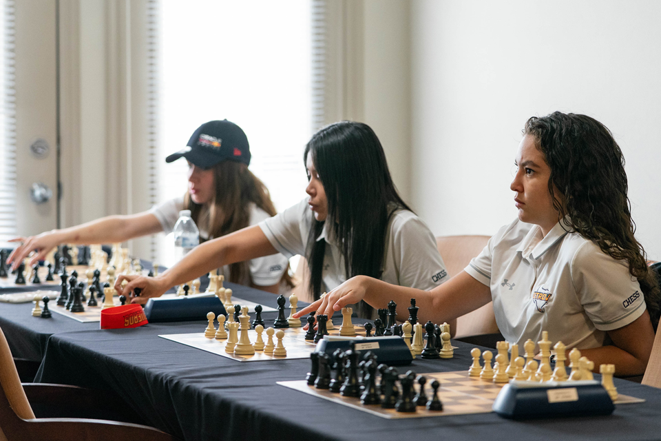 A group of individuals wearing matching light-colored polo shirts sit at a long table, each engaged in a chess game. Multiple chessboards with black and white pieces are arranged in front of them. One person in the foreground extends an arm toward the board to make a move. The setting appears to be a bright indoor room with natural light coming from the left.
