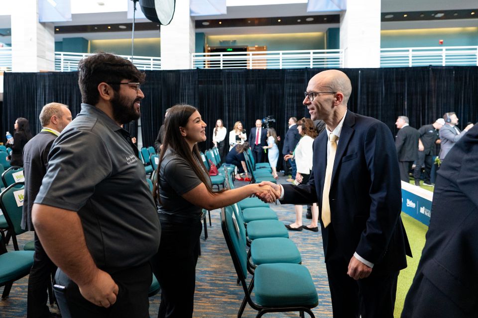 Two attendees shaking hands beside rows of chairs inside an event space, with additional attendees and lighting equipment visible in the background