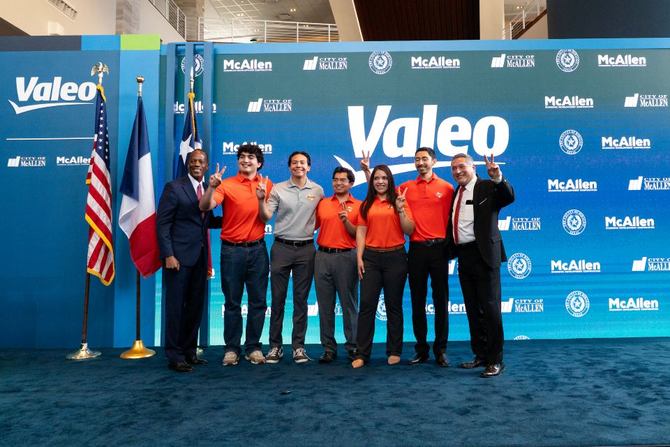 Group of attendees standing on a stage in front of a blue backdrop displaying the Valeo and City of McAllen logos, with United States and Texas flags positioned to the left