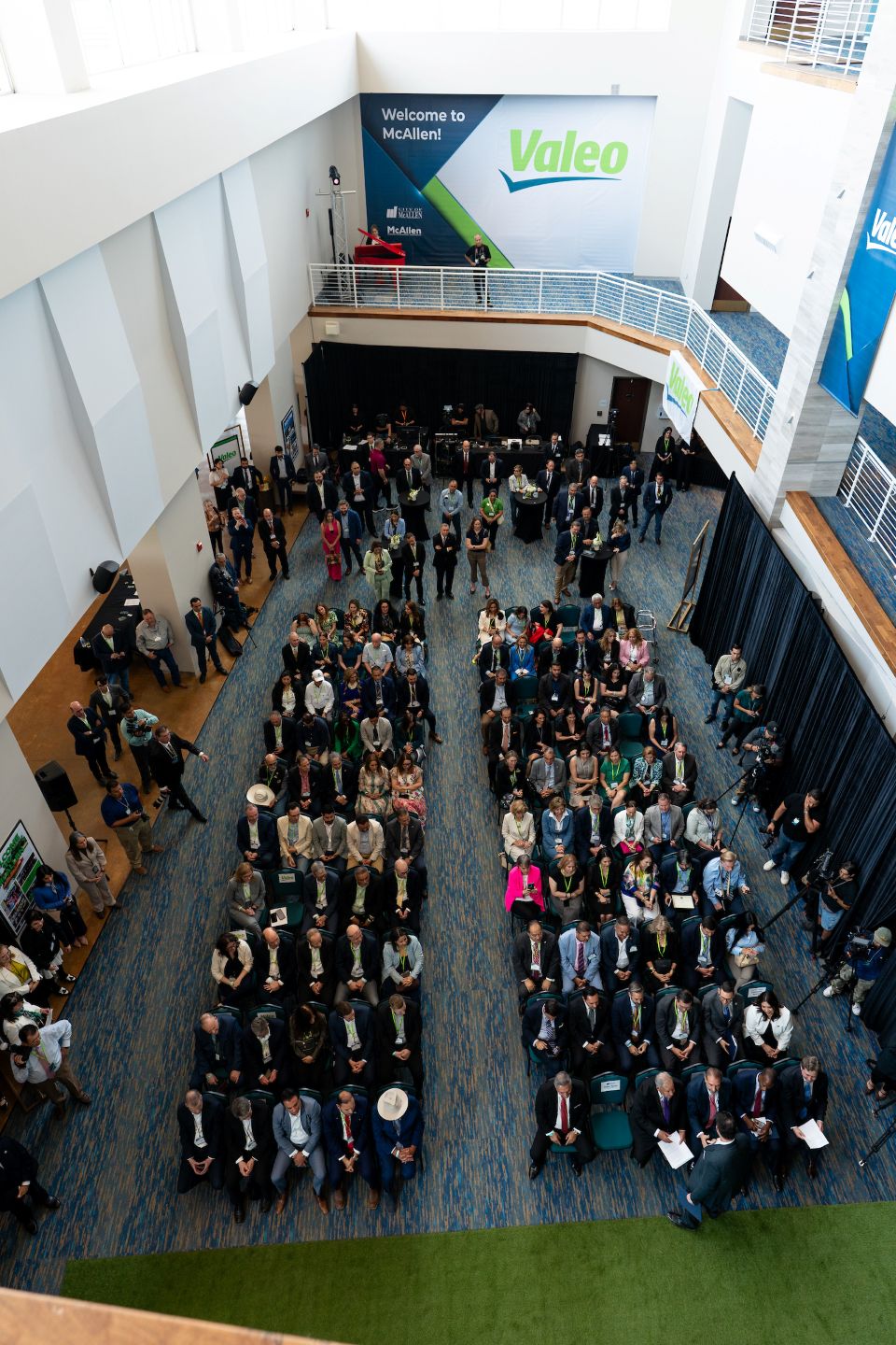 Overhead view of a large seated audience inside a modern atrium during a formal event, with a stage area and a wall sign reading ‘Welcome to McAllen’ and ‘Valeo’ visible in the background